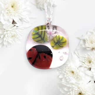 Colorful glass pendant featuring a ladybird, lily pads, and flowers, surrounded by white daisies on a light background.