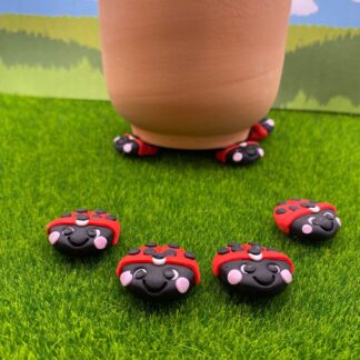 A terracotta pot rests on red and black ladybug-shaped pot feet, with several more ladybug decorations scattered on the artificial grass in front of it.