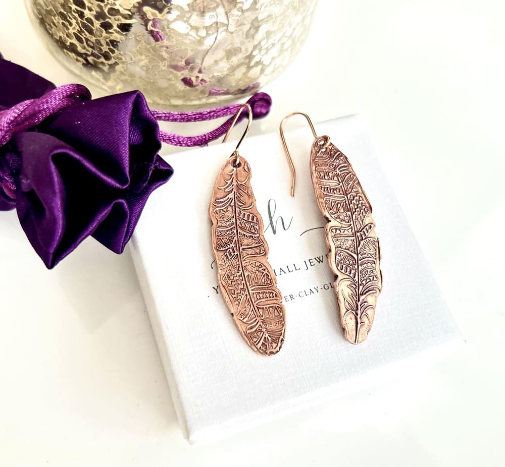 A pair of copper drop earrings in a long textured tribal feather design with a lot of intricate detail and with copper ear wires. They are displayed on top of a white gift box.