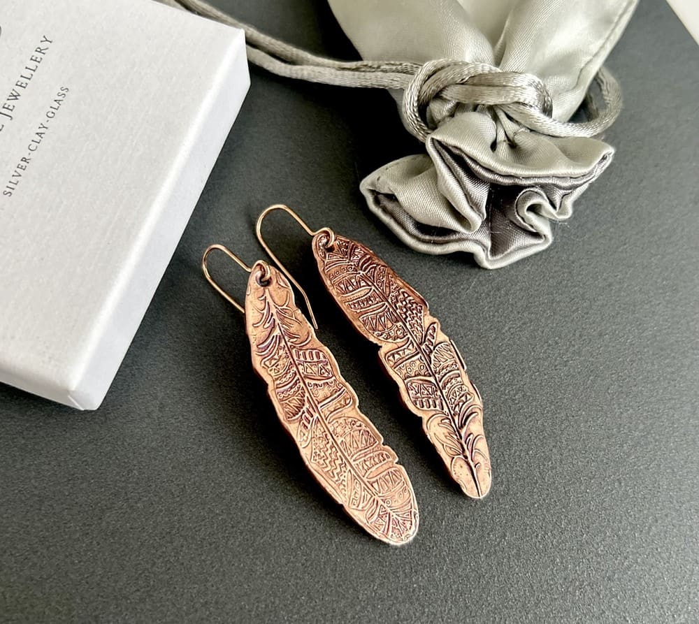 A pair of copper drop earrings in a long textured tribal feather design with a lot of intricate detail and with copper ear wires. They are displayed on a dark surface.