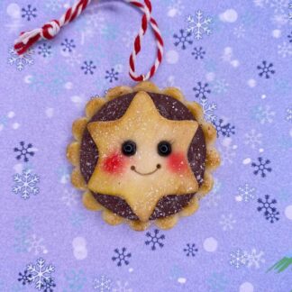 The picture is of a handmade mince pie with a star design on the face, including black bead eyes, a smiling face and rosey cheeks. with a red and white string to hand on the tree. On a lilac background with snowflakes in differing size and colours.