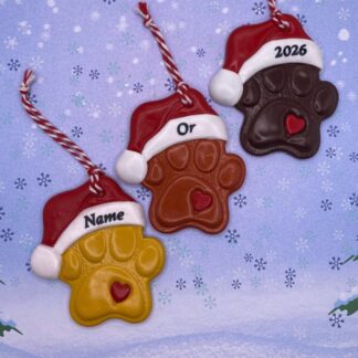 Three paw print Christmas ornaments with Santa hats. One is yellow with "Name" written on the hat, one is brown with "Or" on the hat, and one is dark brown with "2026" on the hat. Each has a small red heart in the center of the paw print.