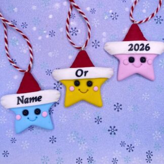 Three star-shaped ornaments with Santa hats are displayed on a blue snowflake background. The ornaments are blue, yellow, and pink, with faces and text. The blue ornament says "Name", the yellow says "Or", and the pink says "2026".