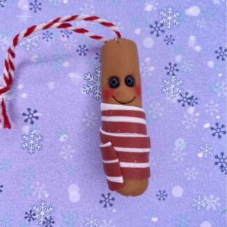 The picture is of a Polymer clay christmas tree decoration - sausage wrapped in bacon also known as - Pigs in blankets. Made with a blend of browns for the sausage and white glitter and a blend of pinks for the bacon. with small black bead eyes, a beaming smile and flushed cheeks all of a lilac backdrop with snowflakes of differing colours and sizes.