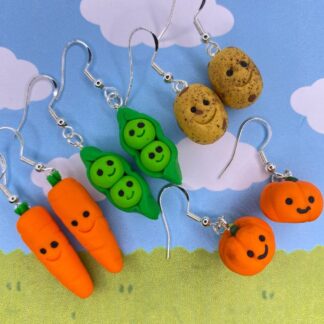 A collection of cute, handmade polymer clay earrings shaped like vegetables, including carrots, peas in a pod, potatoes, and pumpkins, all with smiling faces.
