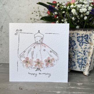 Hand drawn greetings card standing in front of a pot of flowers. Card features a ball gown with applique fabric flowers across the hem. Message reads, happy birthday