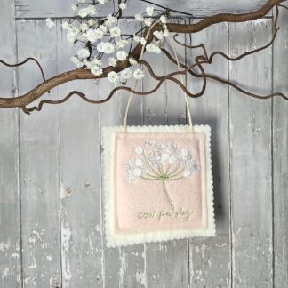 A square felt decoration hangs from a branch. Features an embroidered cow parsley on the front with the words cow parsley embroidered underneath the flower.