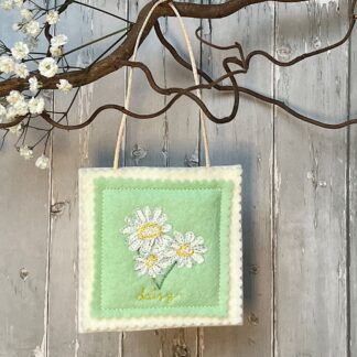 A square green felt decoration hangs from a branch. Features three daisies sewn on front with the word daisy underneath.