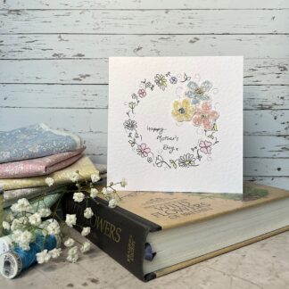 greetings card featuring a wreath of hand drawn flowers and fabric flowers. Message reads happy mothers day. Stands on a book with fabric and flowers nearby.