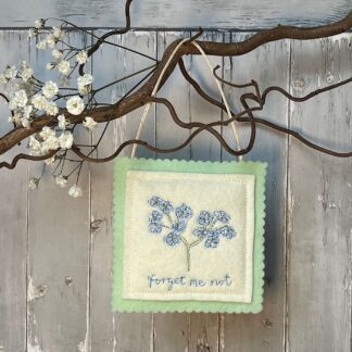 A square felt decoration hanging from a branch. Features blue forget-me-not flowers and the name of the flowers written in embroidery underneath.