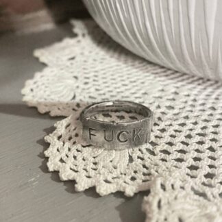 A silver ring with a weathered plate on it, with a text "fuck" stamped on it on irregular letters. The ring lies on top of a neat crocheted tablecloth