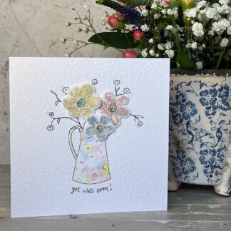 Greetings card with get well soon message with featuring a jug of fabric flowers. Pretty pastel coloured jug, stands in front of a pot of flowers.