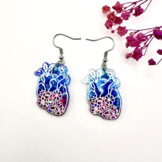 Resin anatomical heart earrings with blue, pink and floral details, displayed on a white background with pink dried flowers. Earrings are hung on surgical steel ear hooks.