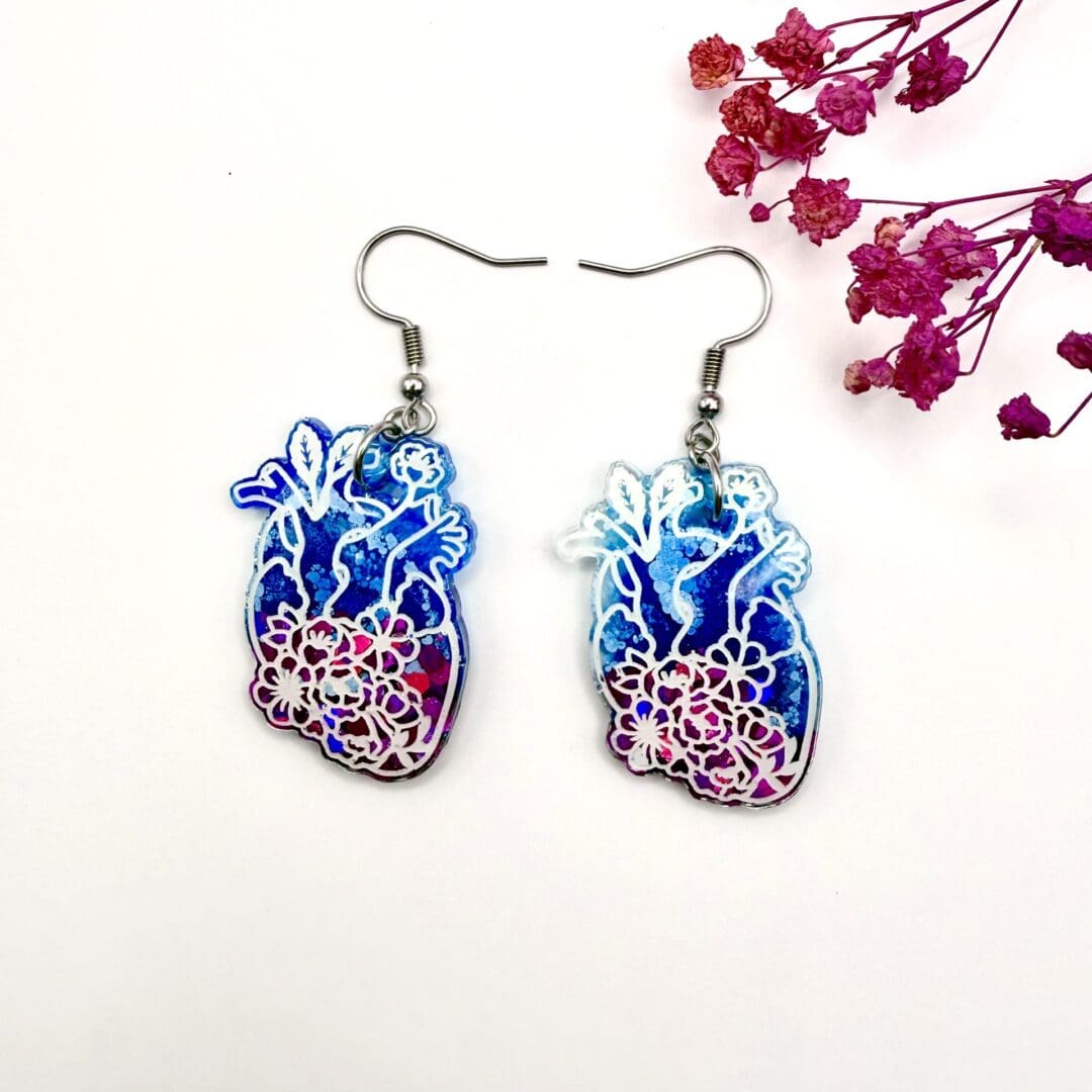 Resin anatomical heart earrings with blue, pink and floral details, displayed on a white background with pink dried flowers. Earrings are hung on surgical steel ear hooks.