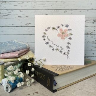 Greetings card featuring a heart of tiny flowers in ink with a fabric flower. Stands on a book with fabric and flowers nearby.