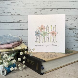 Greetings card of a gate with pastel coloured Liberty fabric flowers attached to it. Message reads congratulations on your new home. Stands on a book with fabric and flowers nearby.