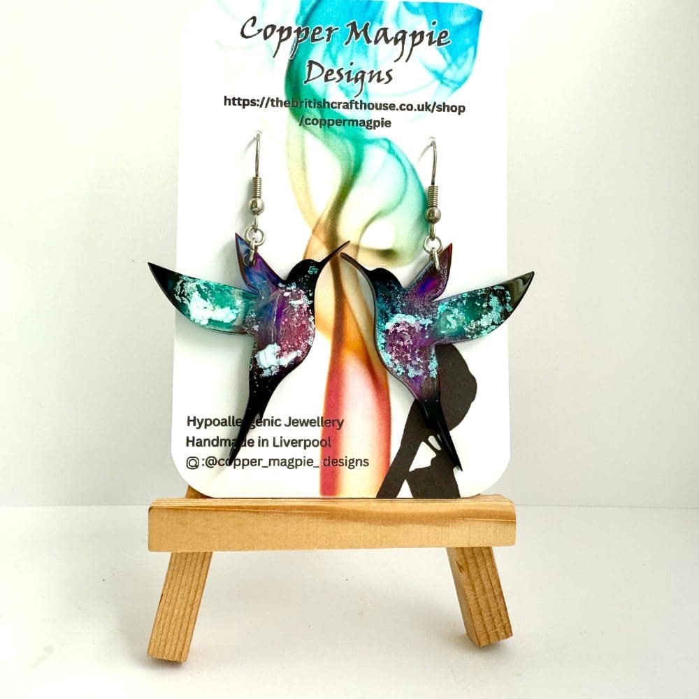 Colourful hummingbird earrings with pink/purple bellies and black tipped green wings. Black tail and head. Hung on surgical steel ear hooks on a branded Copper Magpie Designs card.