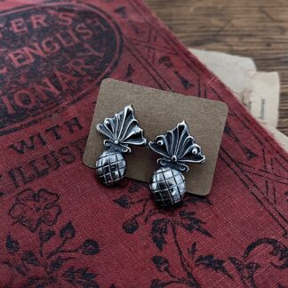 a pair of silver earrings are fastened into a brown piece of cardboard. They resemble pineapples, with a symmetric art-deco style tops and rough handmade fruits with crisscross pattern on them. The pair lie on the cover of an old book, the cover is red with black embossed letters and illustrations.