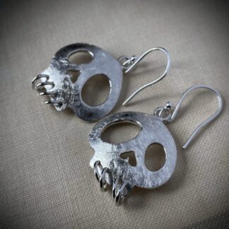 A pair of earrings in silver, shaped like skulls are cut from a sheet of metal. The eyes and nose are hollowed through, and the teeth are dangling silver loops. The surface is scratched so the contrast in between that and the polished edges is accentuated.