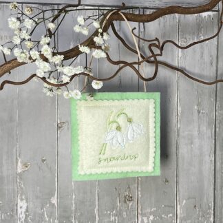 Square felt decoration hangs from a branch with white flowers attached. Decoration features two embroidered snowdrops with the word snowdrop embroidered underneath.