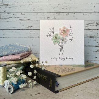 Greetings card featuring a bunch of 3 fabric flowers in pastel colours. Stands on a book with fabric and flowers nearby.