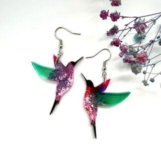 Colourful resin hummingbird earrings with pink and teal wings and dappled bellies with surgical steel earhooks on a white background with flowers
