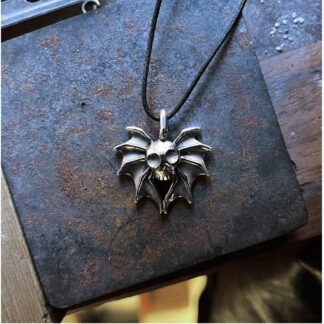 a silver pendant with a crude skull in the middle with bat wings on each side. The metal is very oxidised and it lies on top of a rusty block of metal.
