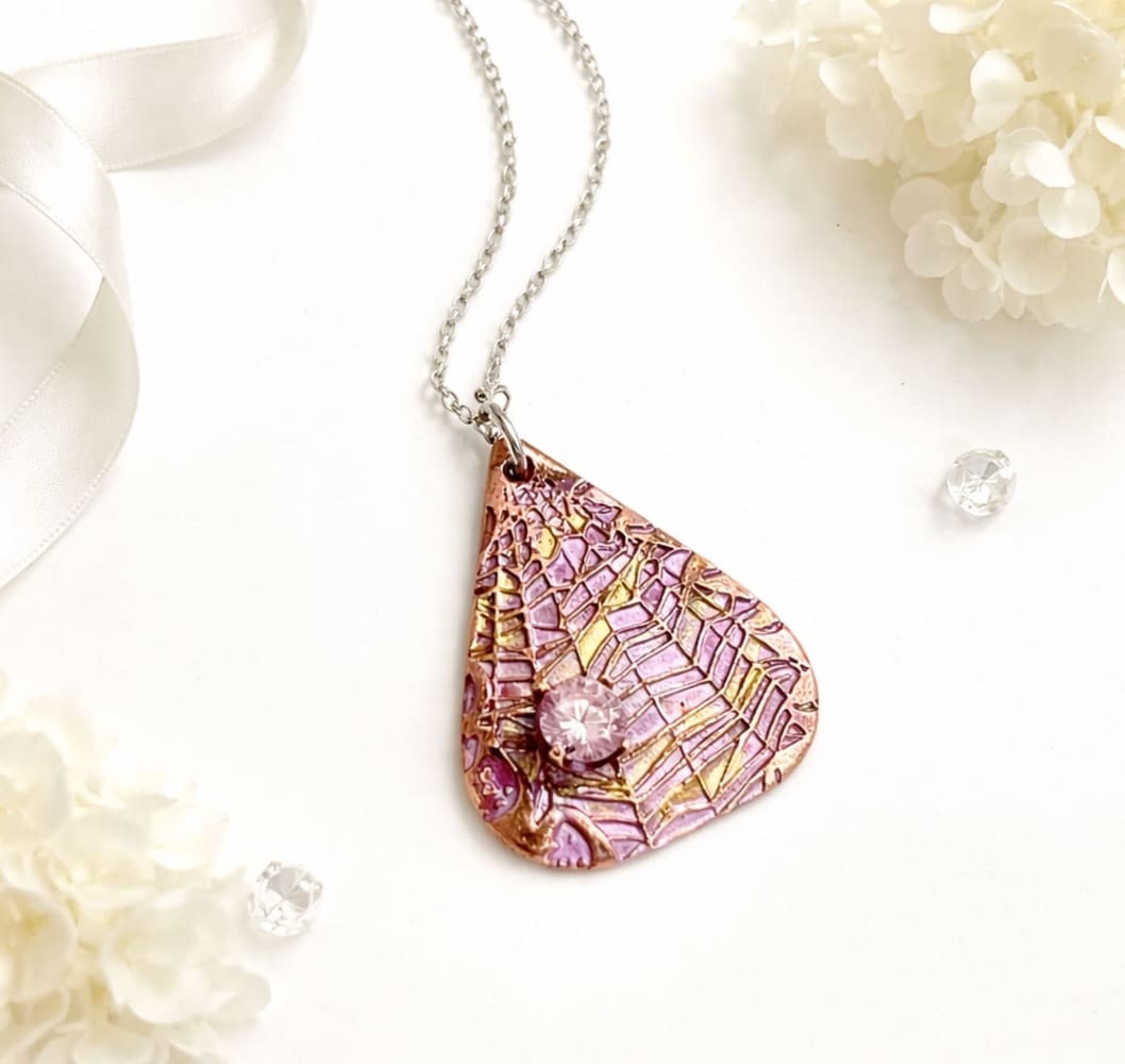 A copper tear drop shaped pendant that has been textured with a cobweb pattern and hand-painted in pink metallic paint, it has a pink cubic zirconia 6mm stone on the lower letf side. It is displayed on a white satin surface.