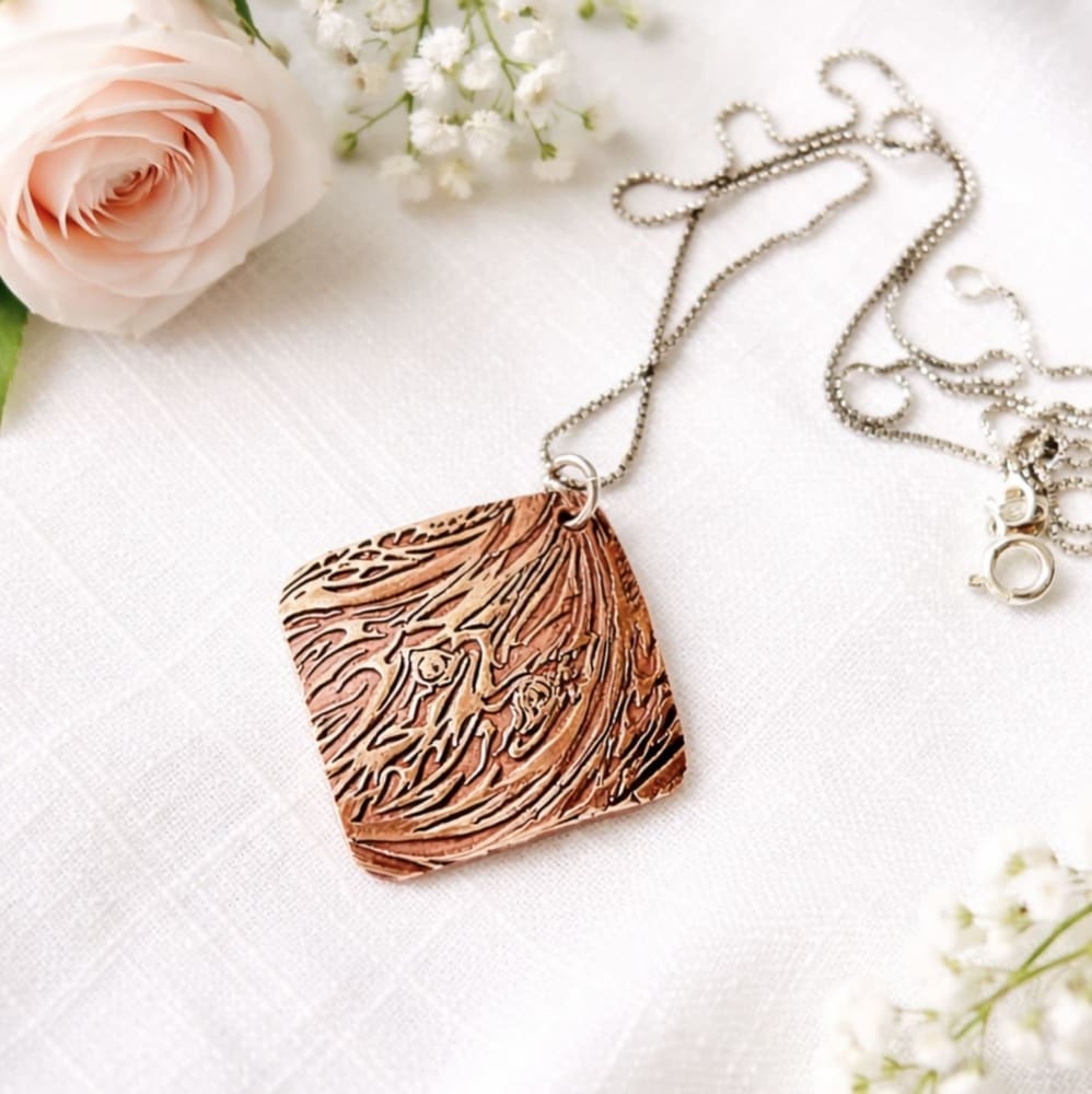A handcrafted textured copper pendant it is in a diamond shape and has a sterling silver chain. The pendant is displayed on a white background with a pink rose and small white flowers.
