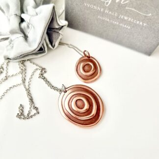 Two copper round pendants with a concentric design, the larger pendant has a sterling silver chain and the small one has a rose gold plated chain. They are displayed on a white background.