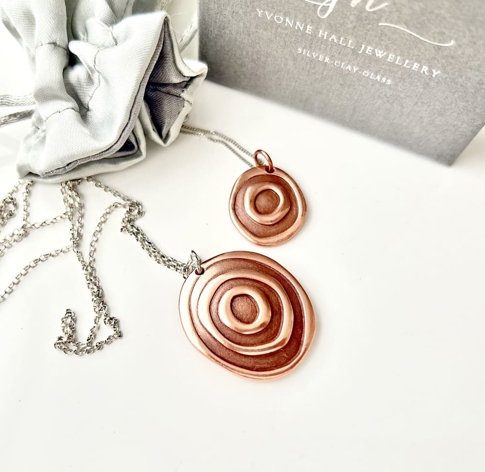 Two copper round pendants with a concentric design, the larger pendant has a sterling silver chain and the small one has a rose gold plated chain. They are displayed on a white background.