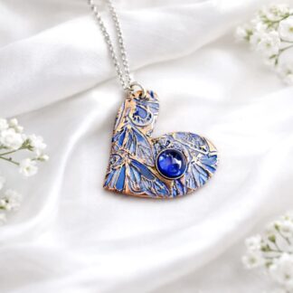 A handcrafted copper heart shaped pendant that is textured and hand-painted with shades if blue, it has a sterling silver chain. It is displayed on a white background.