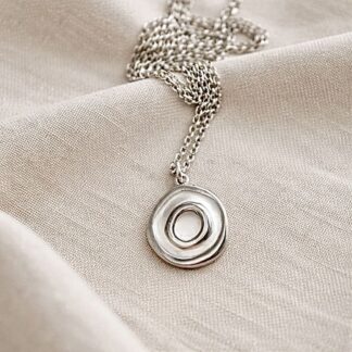 A fine silver small round necklace with a concentric design, it has a sterling silver chain. It is displayed on q beige cloth.
