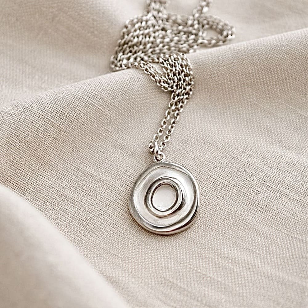 A fine silver small round necklace with a concentric design, it has a sterling silver chain. It is displayed on q beige cloth.