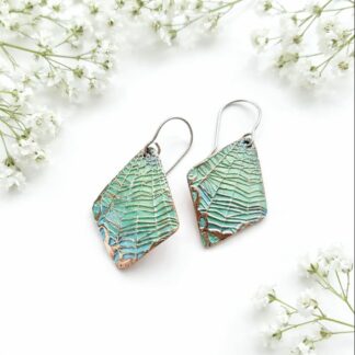 A pair of handcrafted copper earrings with a cob web textures pattern and hand painted in pale turquoise metal paint, in a diamond shape and sterling silver ear wires. They are displayed on a white surface surface with white baby’s breath flowers.
