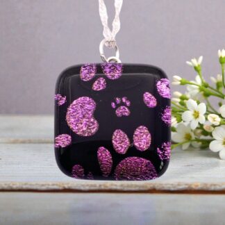 A black pendant featuring sparkling purple paw prints, suspended by a silver chain, against a light background with small white flowers.