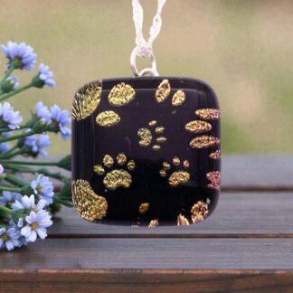A square pendant with black background and shimmering gold paw prints, hanging next to delicate blue flowers on a wooden surface.