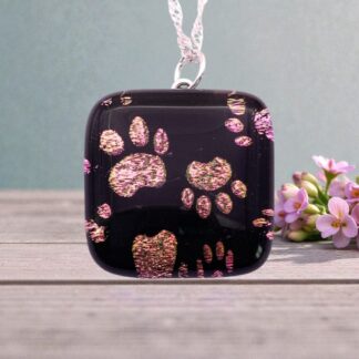 Black pendant with sparkly pink paw prints, hanging on a silver chain, surrounded by delicate flowers on a wooden surface.