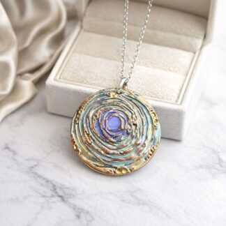 A large round handcraftdd copper pendant, textured with a circle swirl design and hand-painted in pastel greens, with a syerling silver chain. It is displayed leaning on a white jewellery gift box.