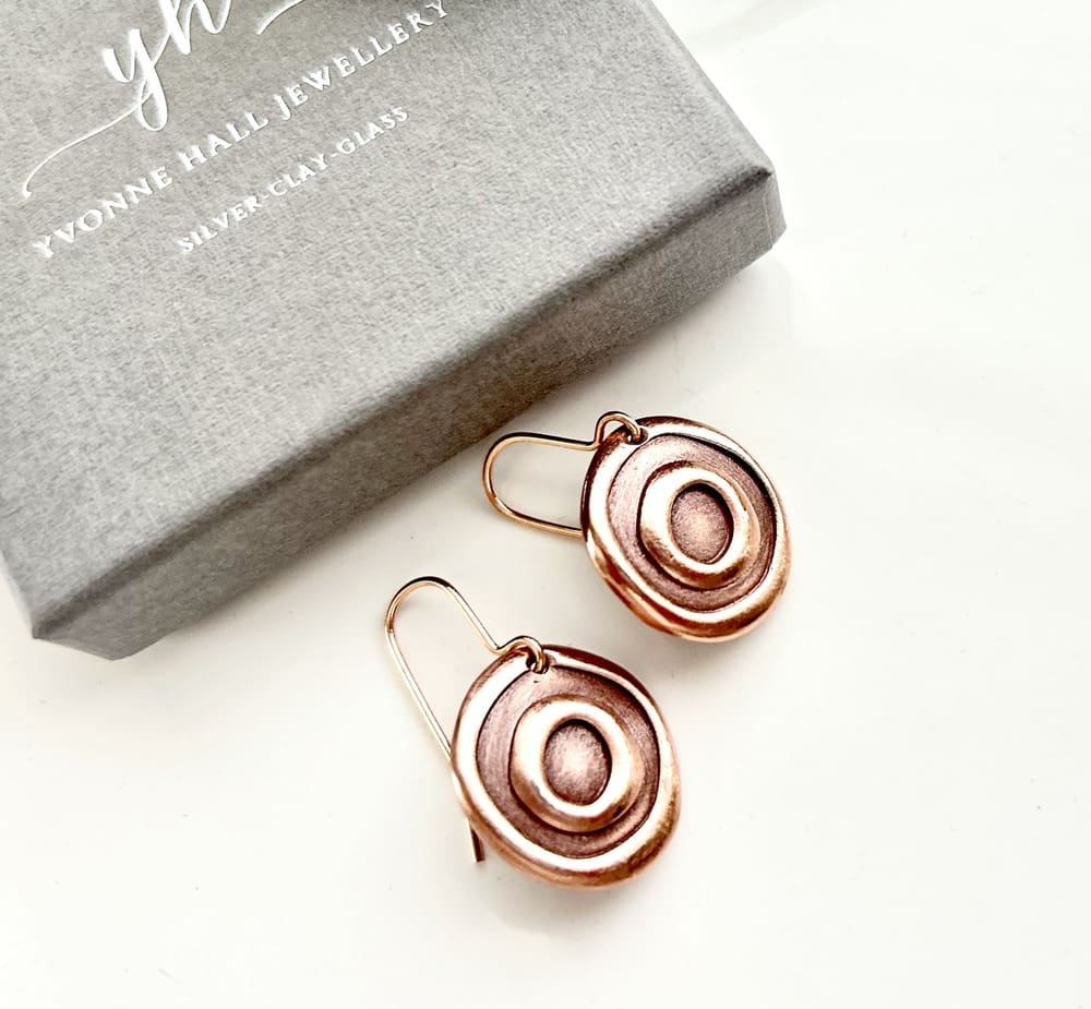 Small round handcrafted copper drop earrings with a raised circle pattern, they have copper ear wires. They are displayed on a white surface with a grey gift box above them.