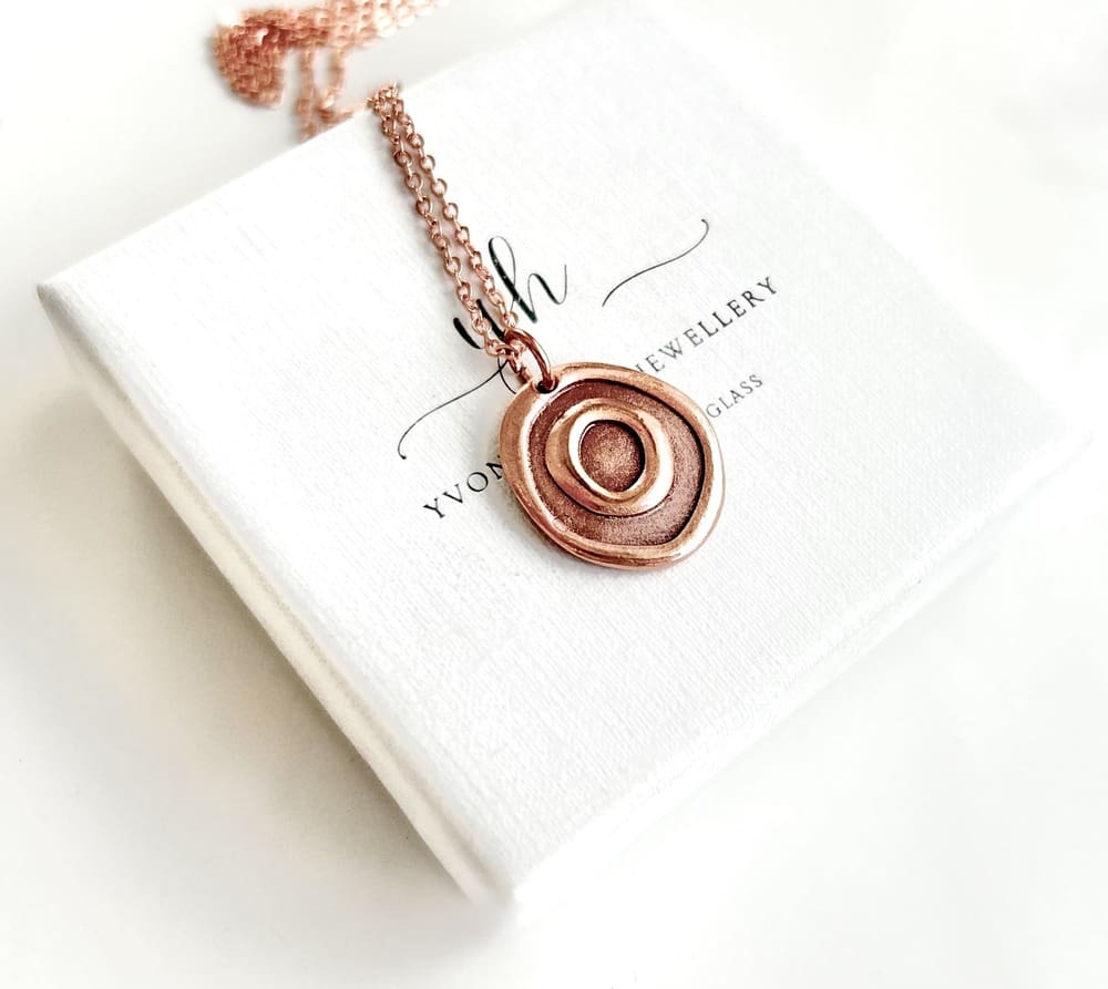 A small copper round pendant with a concentric design, it has a rose gold plated chain. It is displayed on a white gift box set on a white surface.