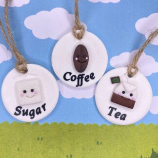 Three round ornaments with cute faces: one with a coffee bean and the word "Coffee", one with a sugar cube and the word "Sugar", and one with a teabag and the word "Tea".