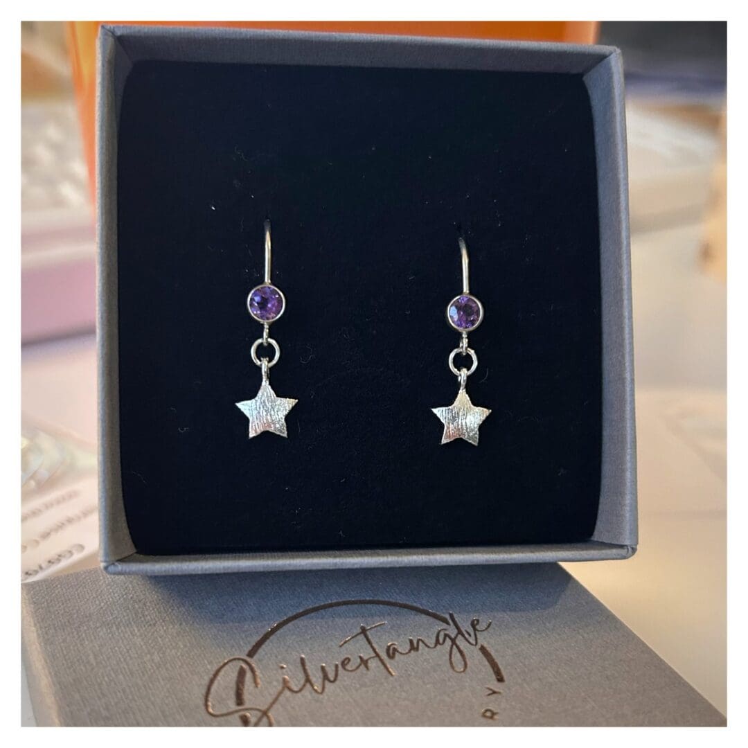 Small silver star hangs from a silver ear wire in which is set a round amethyst gemstone. The earrings are in a grey box with black foam insert.