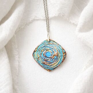 A copper handcrafted pendant in a softened triangle shape with a circular textured and hand-painted in turquoise and green metallic paints, with a sterling silver bail and chain. It is displayed on a white background.