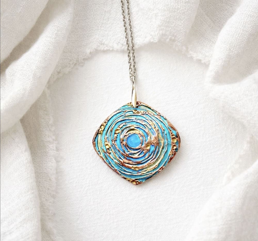 A copper handcrafted pendant in a softened triangle shape with a circular textured and hand-painted in turquoise and green metallic paints, with a sterling silver bail and chain. It is displayed on a white background.