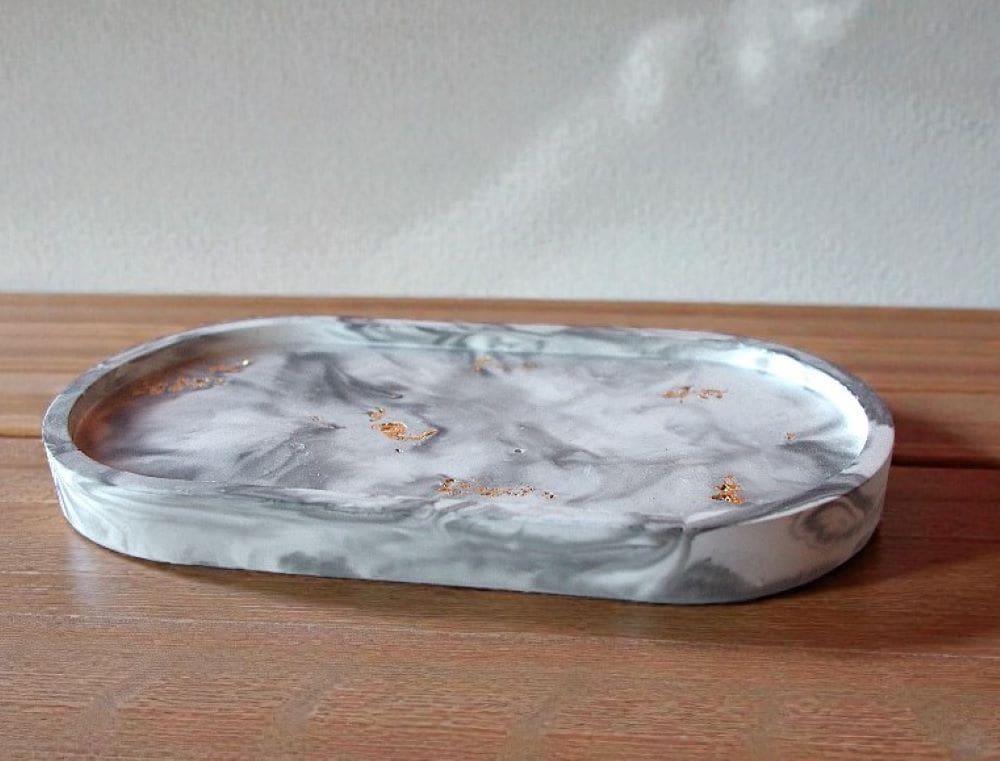 An oval trinket tray is sitting on top of a light wooden counter, in front of an off white wall. The tray has a black and white marble pattern, set with gold flecks.