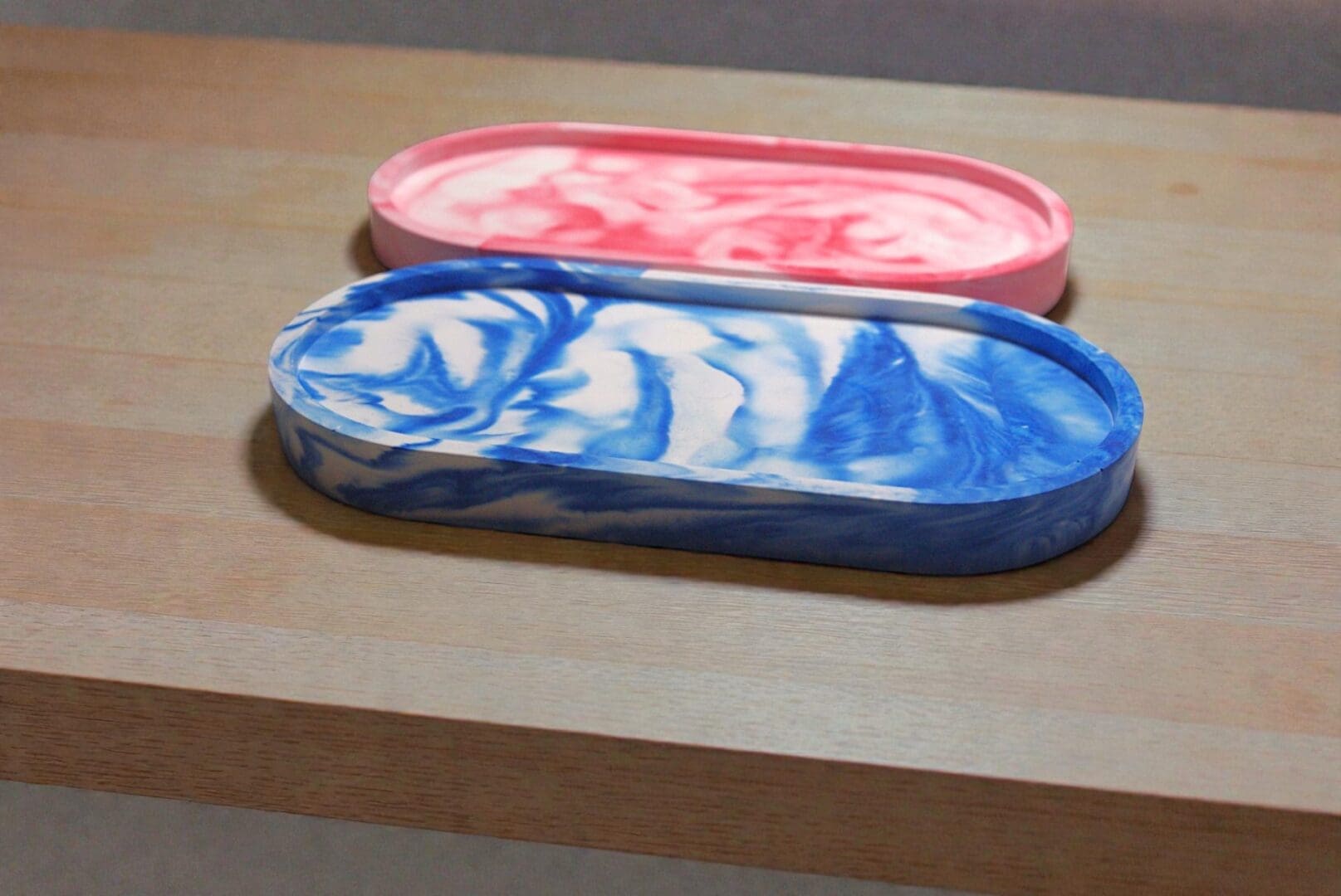 Two oval trinket trays next to each other on a light wooden countertop. The tray nearest to the front is blue marble, the one next to it is red marble.