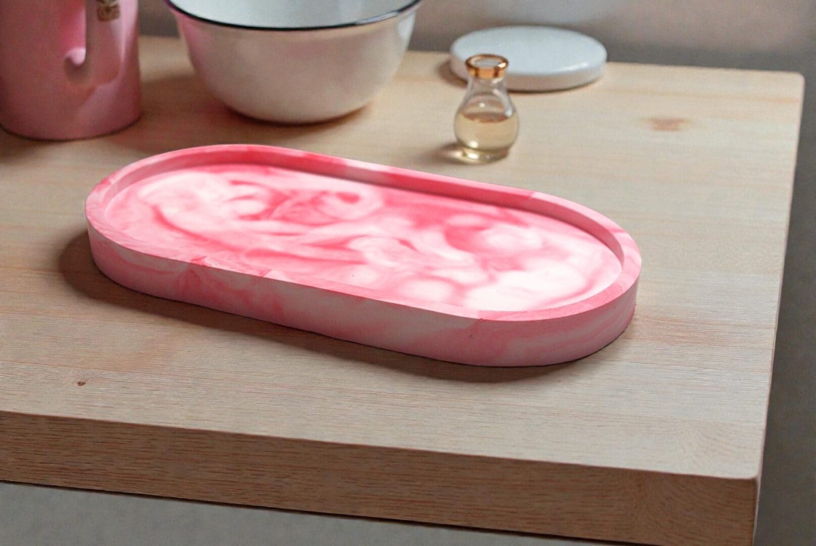 A red and white marble effect trinket tray, sits on a light wood countertop in front of a small perfume bottle, a pink mug and and a white bowl.