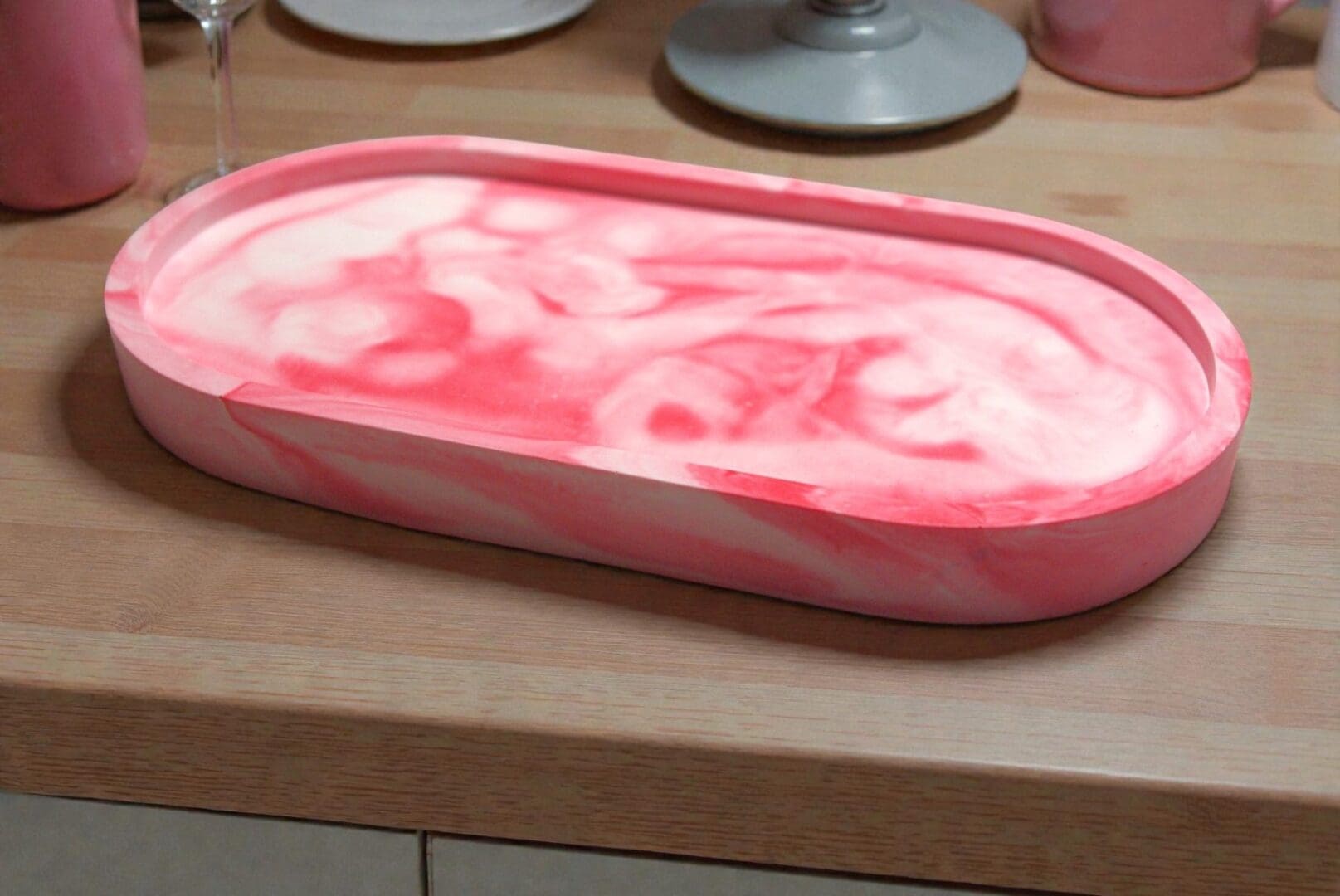 An oval, red and white trinket tray sits on a pale wooden countertop. Behind the tray are the bottoms of three glasses.