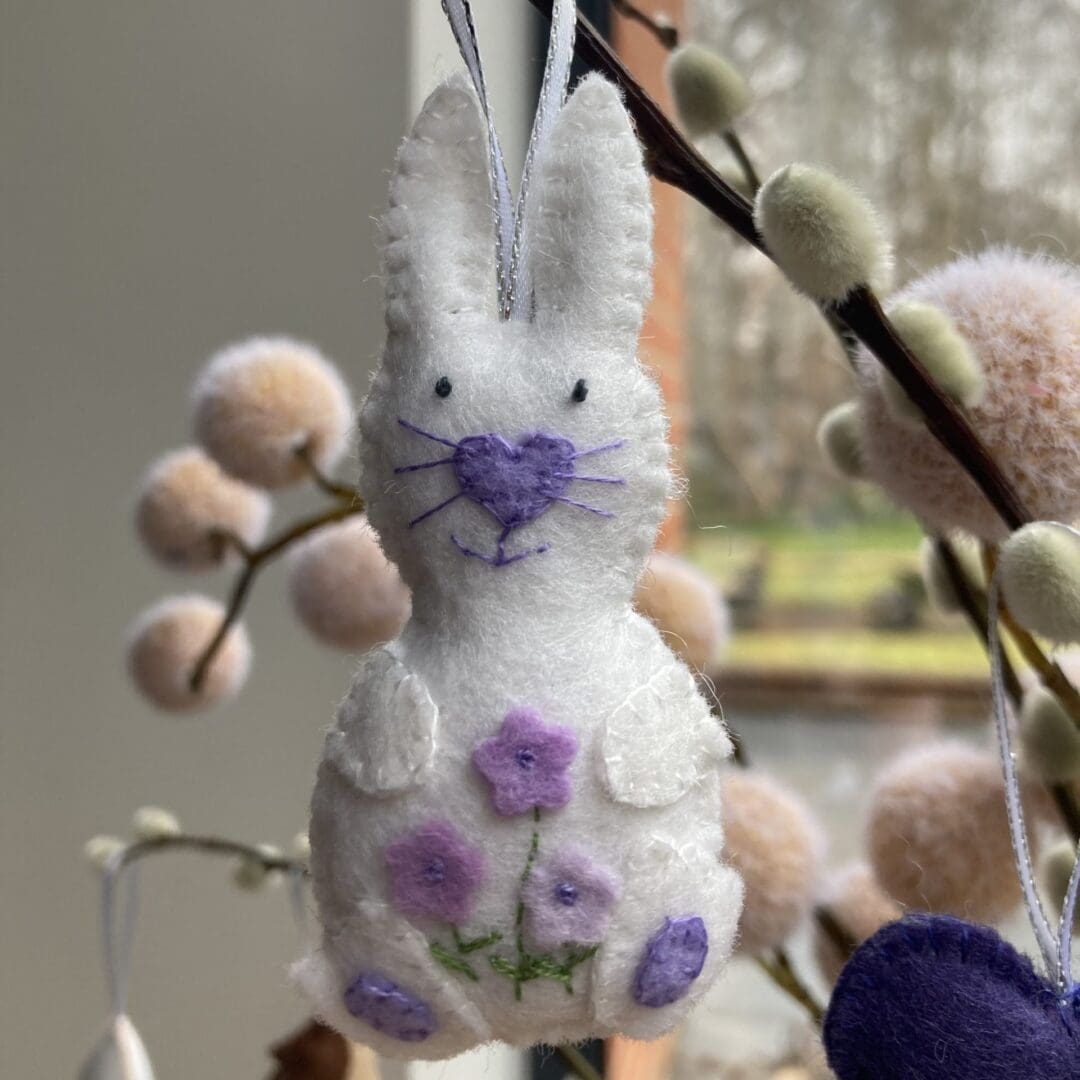 Handmade white felt bunny carrying purple flowers and has purple nose, whiskers and paws